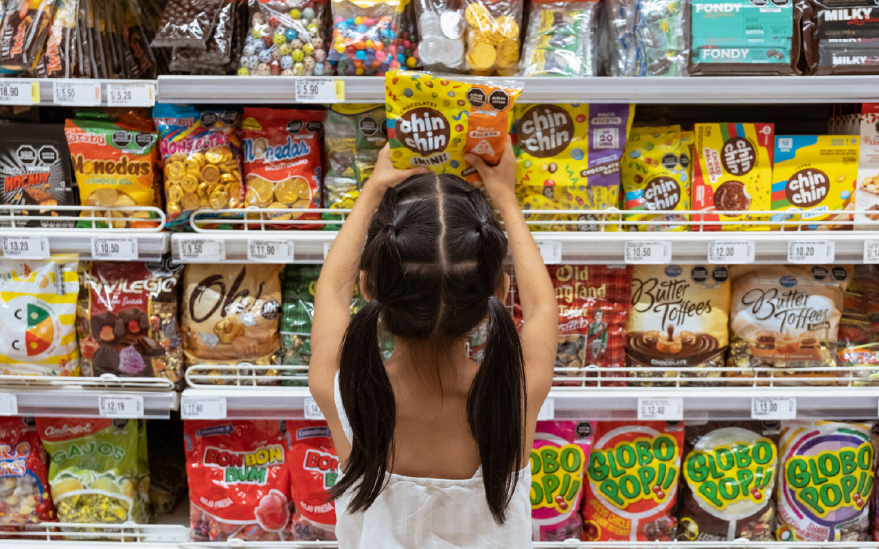 Niña de espaldas y frente a un estante de supermercado repleto de dulces envueltos en envases de plástico colores