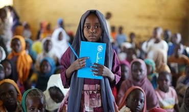 niña al frente con una carpeta de UNICEF en Sudán, rodeada de muchos más niños y niñas