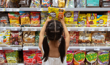 Niña de espaldas y frente a un estante de supermercado repleto de dulces envueltos en envases de plástico colores