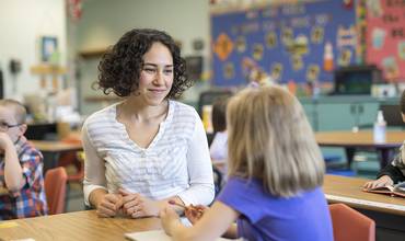 Una maestra habla con una niña en una mesa de un aula de educación infantil