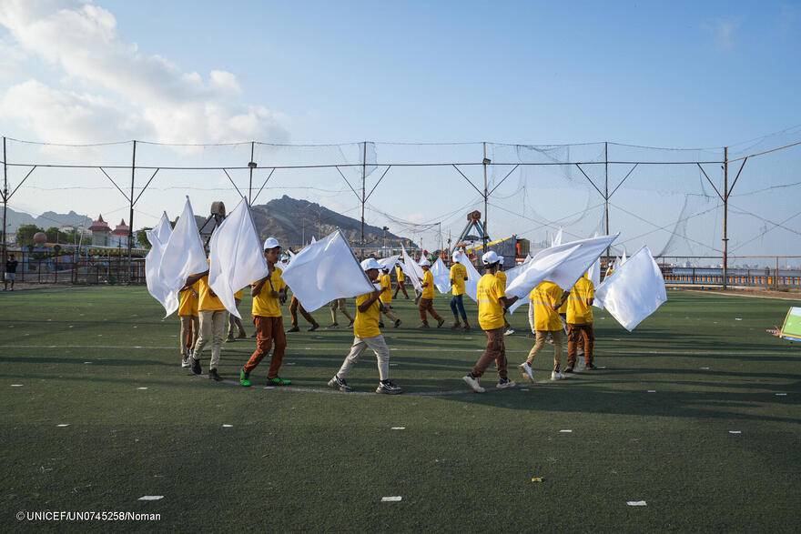 Niños y niñas forman un círculo mientras ondean banderas blancas, sobre el césped del estadio Fun City de Aden, en Yemen.