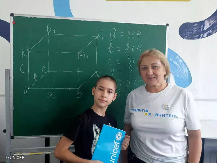 Denys posa delante de la pizarra con su profe de mates y el material escolar de UNICEF. Denys con su profesora