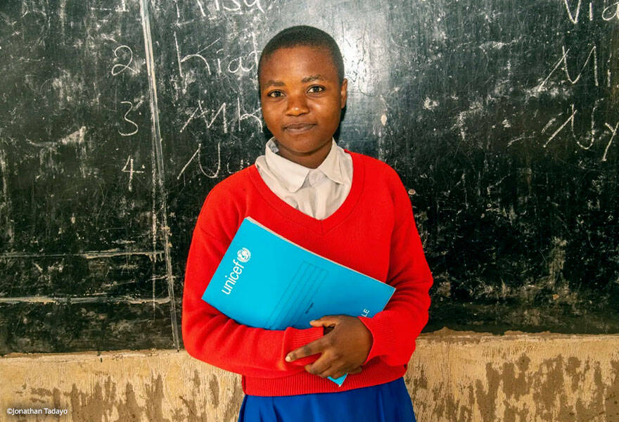 Unidos Por - Educación Vestina, feliz delante de la pizarra de clase con el material escolar proporcionado por UNICEF.